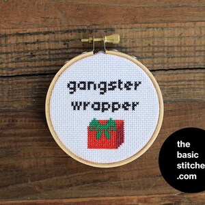 Peut inclure: Un cercle de broderie blanc avec le texte "gangster wrapper" et un cadeau rouge et vert avec un nœud. Le cercle est sur une surface en bois.