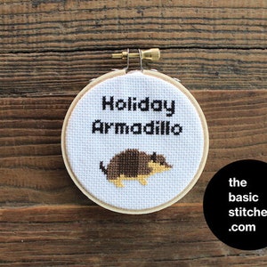 May include: A white embroidery hoop with the text "Holiday Armadillo" and a brown and yellow embroidered armadillo. The hoop is on a wooden surface. The text "the basic stitches .com" is in a black circle below the hoop.