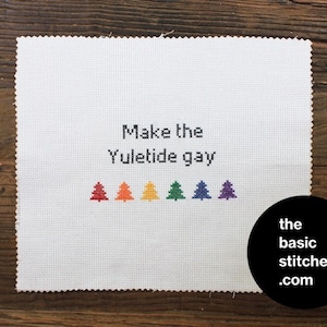 May include: A white cross-stitch piece with the phrase "Make the Yuletide gay" stitched in black. Below the text are six colourful cross-stitched Christmas trees in red, orange, yellow, green, blue, and purple. A black circle with white text reads "the basic stitches .com".