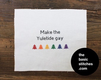 Puede incluir: Tela blanca bordada a punto de cruz con el texto "Make the Yuletide gay" en negro. Debajo, seis árboles de Navidad coloridos bordados en rojo, naranja, amarillo, verde, azul y morado. Un círculo negro con texto blanco dice "the basic stitches .com".