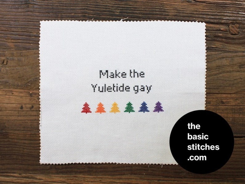 May include: A white cross-stitch piece with the phrase "Make the Yuletide gay" stitched in black. Below the text are six colourful cross-stitched Christmas trees in red, orange, yellow, green, blue, and purple. A black circle with white text reads "the basic stitches .com".