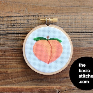 May include: A peach cross-stitch embroidery pattern in a round wooden hoop. The peach is a light orange color with a green leaf on top. The background is white.