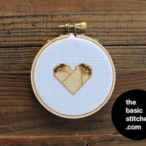 May include: A white embroidery hoop with a cross-stitch design of two burritos forming a heart shape. The burritos are filled with brown beans and green vegetables. The hoop is on a wooden surface. The text "the basic stitches.com" is on a black circle in the lower right corner of the image.