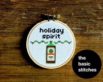 Puede incluir: Un aro de bordado blanco con un diseño bordado en verde y rojo de una botella de licor con el texto "holiday spirit".