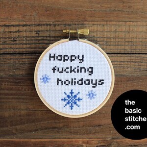 以下が含まれることがあります： 白い刺繍枠に、黒い文字で「Happy fucking holidays」と書いてあり、青い雪の結晶のデザインが施されています。枠は木製の表面の上に置かれています。
