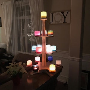 May include: A wooden candle holder shaped like a Christmas tree with multiple tiers. The tiers are filled with colorful glass candle holders with lit candles. The word "JOY" is on a sign on the wall behind the candle holder.