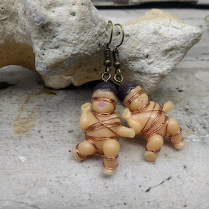 May include: A pair of earrings featuring small plastic baby dolls wrapped in copper wire. The dolls have brown hair, closed eyes, and pink lips. The earrings are attached to bronze-colored hooks.