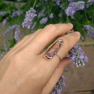 May include: A rose gold ring with a raw, pink and brown crystal set in the band. The ring is on a hand with lavender flowers in the background.