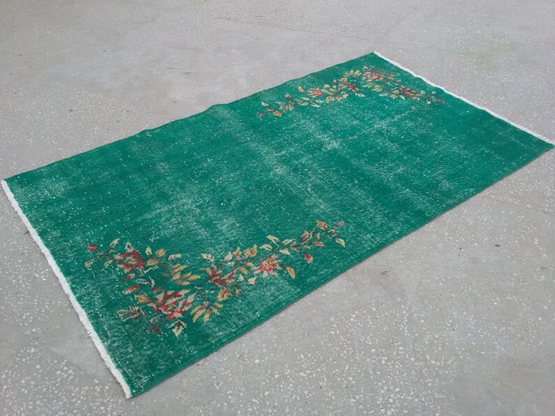 Pode incluir: Um tapete verde vintage com um padr&atilde;o floral em tons de vermelho, amarelo e branco. O tapete tem um aspecto desgastado e &eacute; franjado em todos os lados.