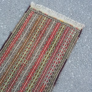 May include: A long, narrow rug with a geometric pattern in shades of red, green, brown, and white. The rug has a fringe on both ends.