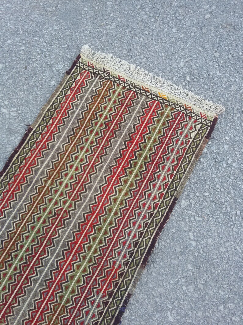 May include: A long, narrow rug with a geometric pattern in shades of red, green, brown, and white. The rug has a fringe on both ends.