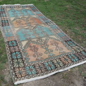 Puede incluir: Una alfombra turca vintage con un patr&oacute;n geom&eacute;trico en tonos azules, marrones y beige. La alfombra tiene un borde con flecos y est&aacute; colocada sobre una superficie de hierba.