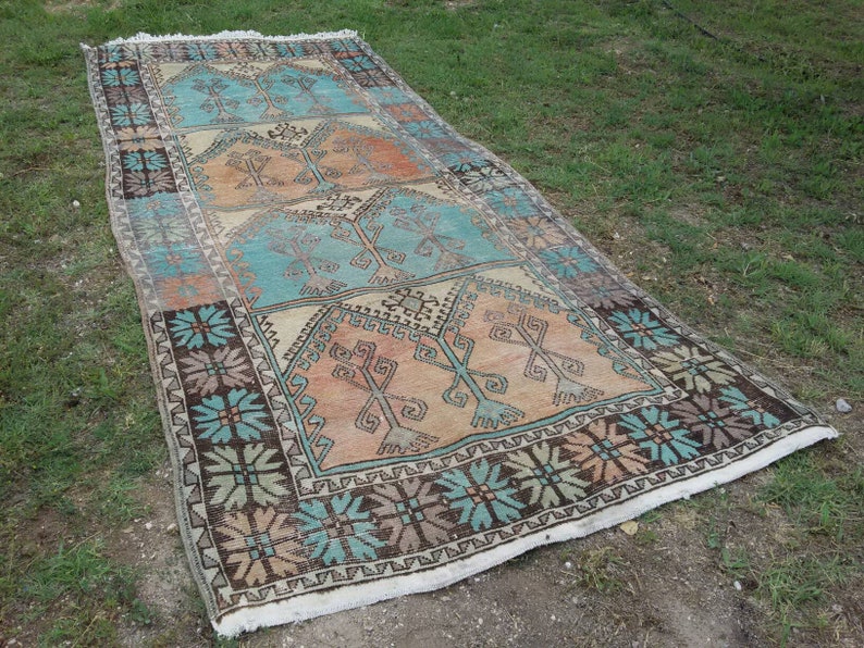 Puede incluir: Una alfombra turca vintage con un patr&oacute;n geom&eacute;trico en tonos azules, marrones y beige. La alfombra tiene un borde con flecos y est&aacute; colocada sobre una superficie de hierba.