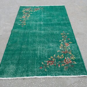 Pode incluir: Um tapete verde vintage com um padr&atilde;o floral em tons de amarelo, laranja e vermelho. O tapete tem um aspecto desgastado e uma borda branca com franjas.