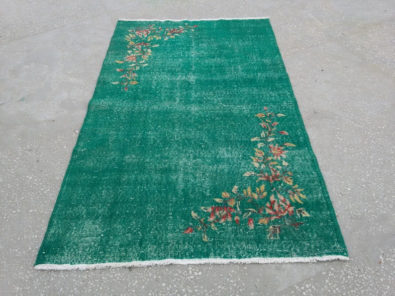 Pode incluir: Um tapete verde vintage com um padr&atilde;o floral em tons de amarelo, laranja e vermelho. O tapete tem um aspecto desgastado e uma borda branca com franjas.