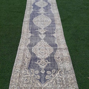 May include: A vintage runner rug with a faded blue background and intricate white floral patterns. The rug has a fringed edge and is laid out on a green surface.