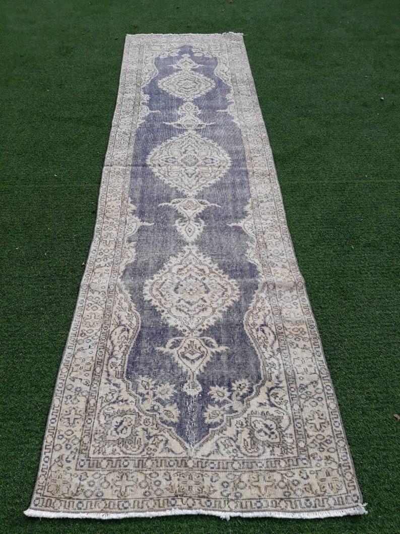 May include: A vintage runner rug with a faded blue background and intricate white floral patterns. The rug has a fringed edge and is laid out on a green surface.