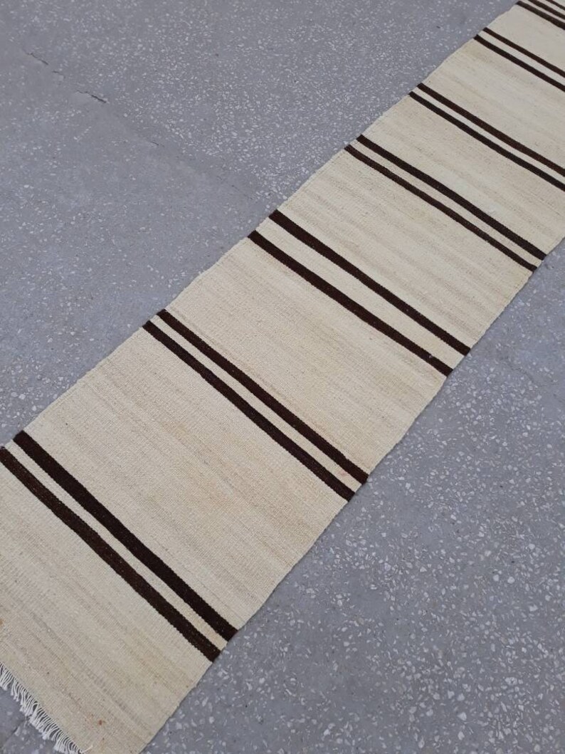 May include: A white and brown striped runner rug with fringe on the ends. The rug is made from a natural fibre and has a simple, classic design.