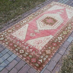 May include: A vintage Turkish rug with a pink and brown color scheme. The rug has a geometric pattern with a central medallion. The rug is laid out on a brick walkway.