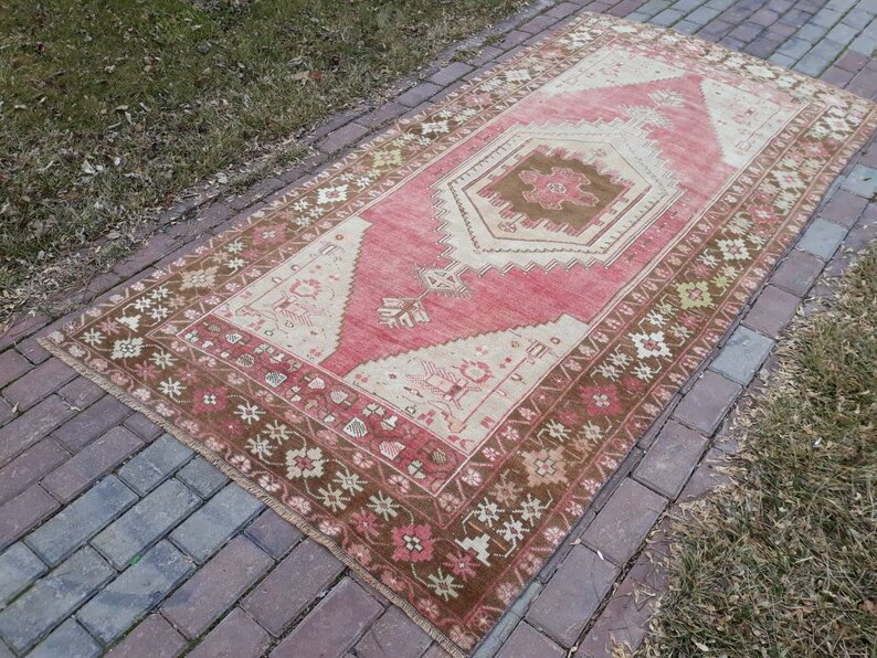 May include: A vintage Turkish rug with a pink and brown color scheme. The rug has a geometric pattern with a central medallion. The rug is laid out on a brick walkway.