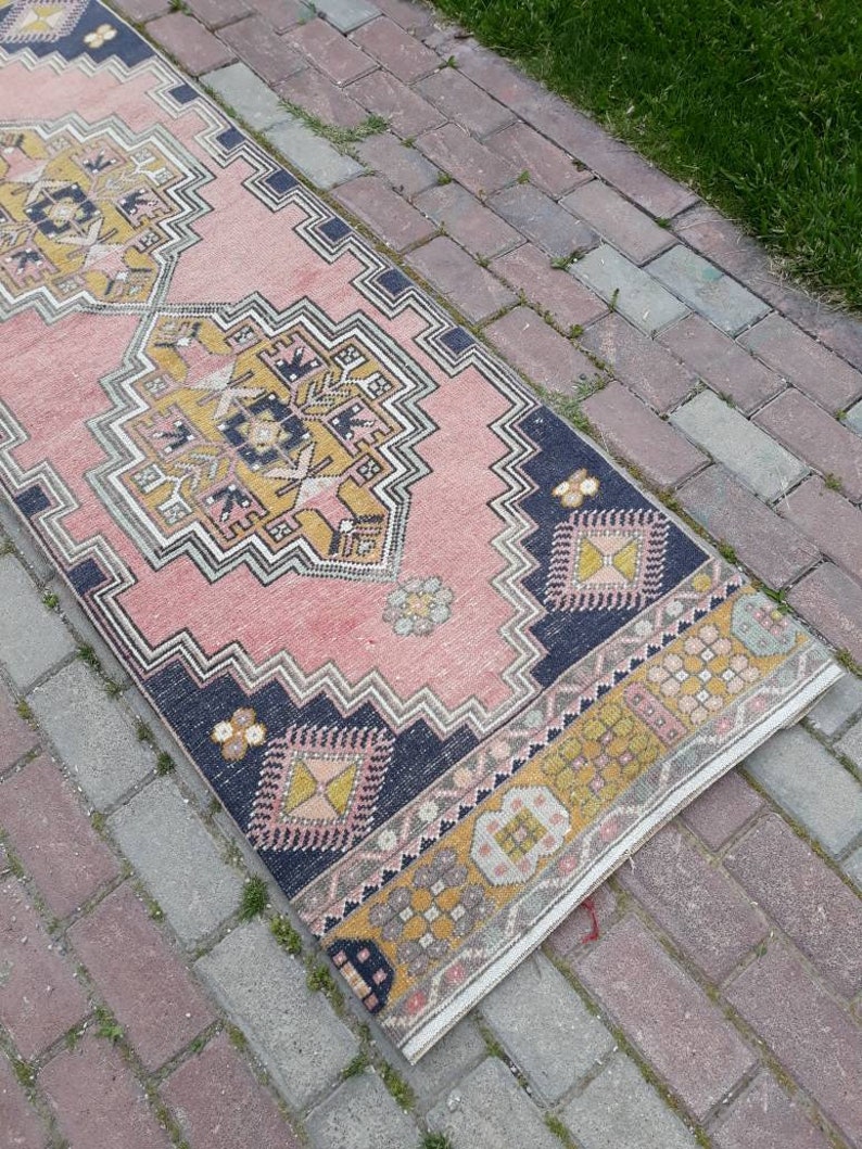 May include: A pink and yellow patterned runner rug with a dark blue border. The rug is laid out on a brick walkway.