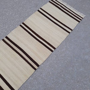 May include: A long, narrow, cream-coloured rug with brown stripes. The rug has a fringe on the edges.