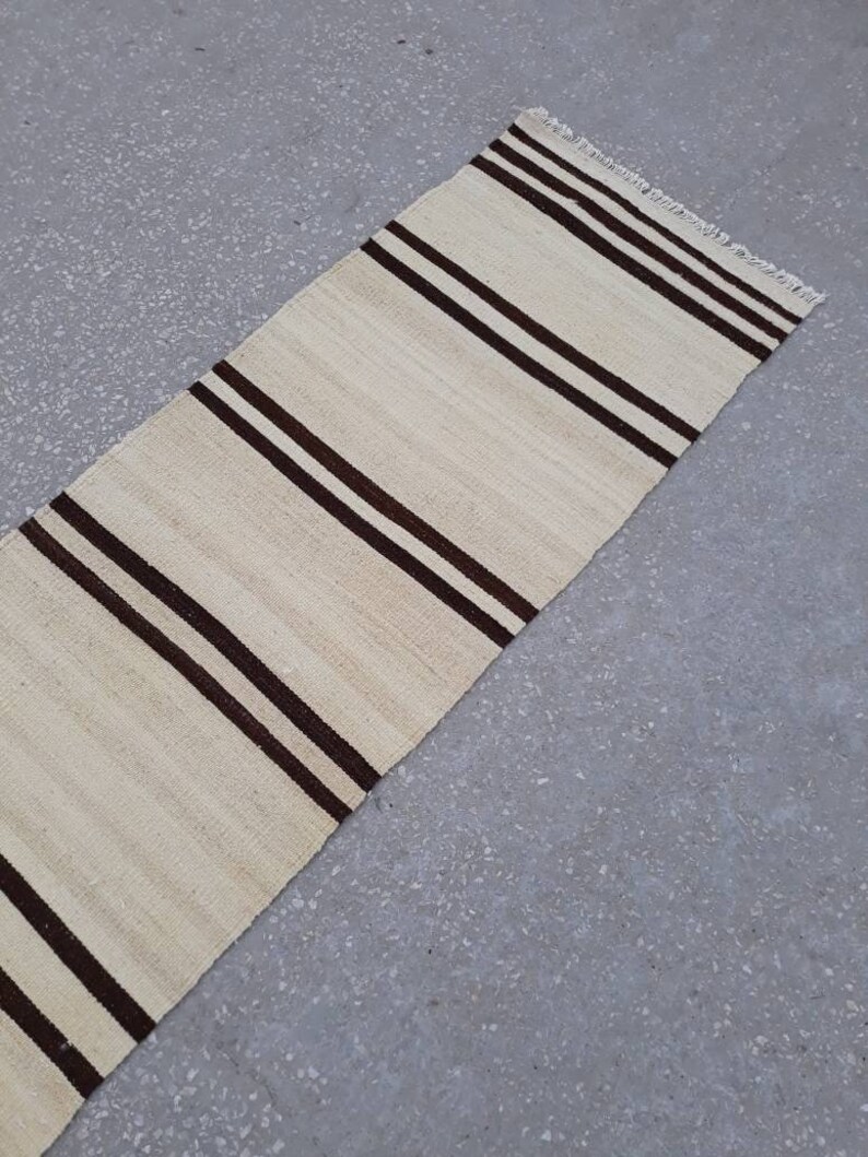 May include: A long, narrow, cream-coloured rug with brown stripes. The rug has a fringe on the edges.