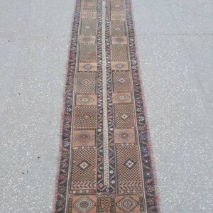 May include: A long, narrow rug with a traditional Persian design. The rug is primarily beige with brown, red, and black accents. The rug has a fringe on both ends.