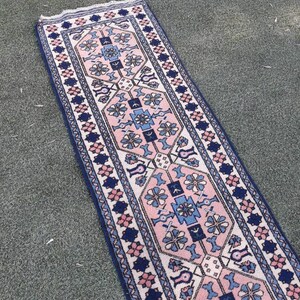 May include: A long, narrow rug with a traditional Persian design. The rug has a white background with pink, blue, and navy blue geometric patterns. The rug is fringed on the ends.