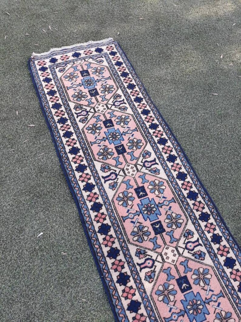 May include: A long, narrow rug with a traditional Persian design. The rug has a white background with pink, blue, and navy blue geometric patterns. The rug is fringed on the ends.