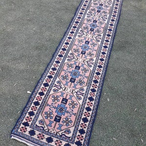 May include: A long, narrow rug with a traditional Persian design. The rug has a cream background with a central pattern of pink, blue, and white flowers and geometric shapes. The border is a dark blue with a repeating pattern of pink and white flowers.
