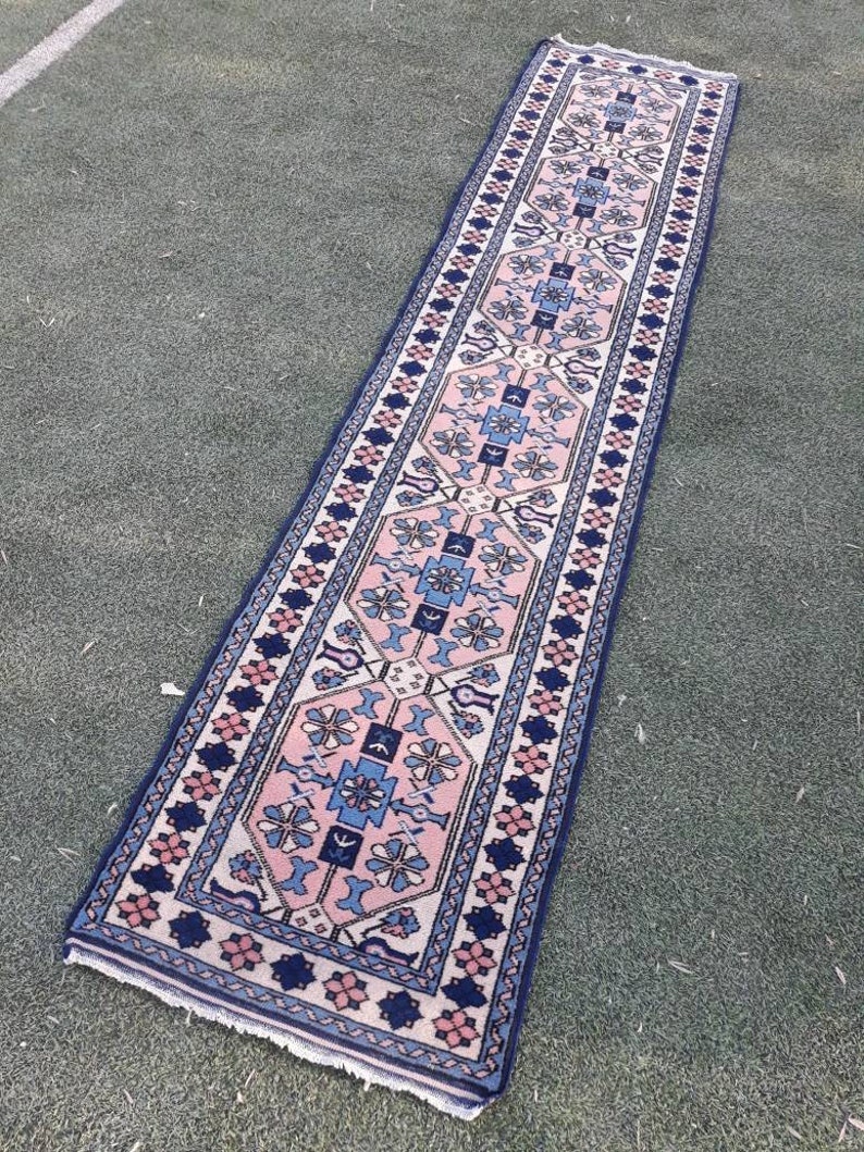 May include: A long, narrow rug with a traditional Persian design. The rug has a cream background with a central pattern of pink, blue, and white flowers and geometric shapes. The border is a dark blue with a repeating pattern of pink and white flowers.