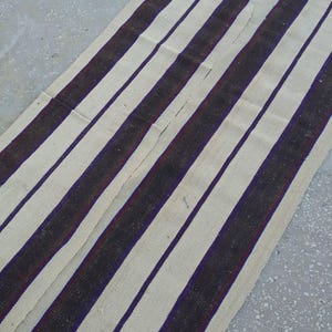 May include: A white and black striped rug with a subtle purple stripe. The rug is made of a woven fabric and has a rustic, vintage look.