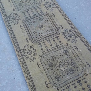May include: A beige runner rug with a geometric pattern in black. The rug has a faded look and is made of a woven material.