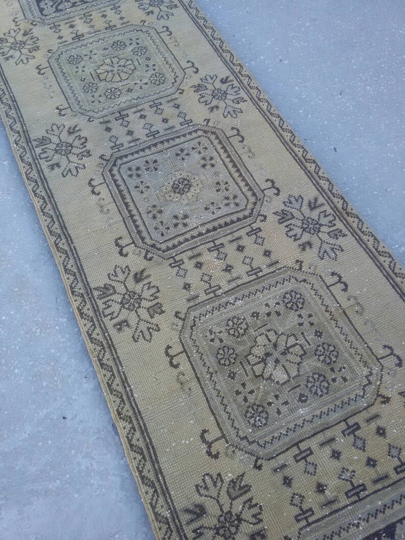 May include: A beige runner rug with a geometric pattern in black. The rug has a faded look and is made of a woven material.