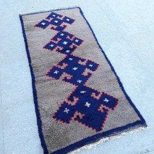 May include: Rectangular rug with a gray background and a central design in blue, red, and white. The design is a repeating geometric pattern. The rug has a dark blue border and a fringed edge.
