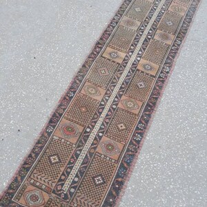 May include: A long, narrow rug with a traditional Persian design. The rug is woven with a variety of colors, including red, brown, and cream. The rug has a fringe on both ends.