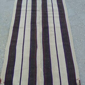 May include: A long, narrow, cream-colored rug with alternating black and purple stripes. The rug is woven with a simple, rustic design.