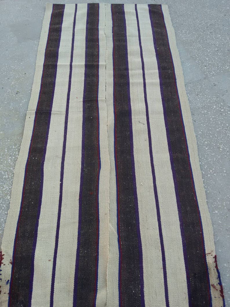 May include: A long, narrow, cream-colored rug with alternating black and purple stripes. The rug is woven with a simple, rustic design.