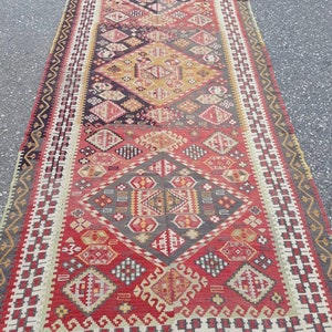 May include: A vintage Turkish rug with a geometric pattern in shades of red, yellow, black, and white. The rug has a fringe border and is woven with intricate details.