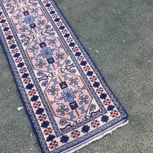 May include: A long, narrow rug with a pink, blue, and white geometric pattern. The rug has a fringed edge and is made of wool.