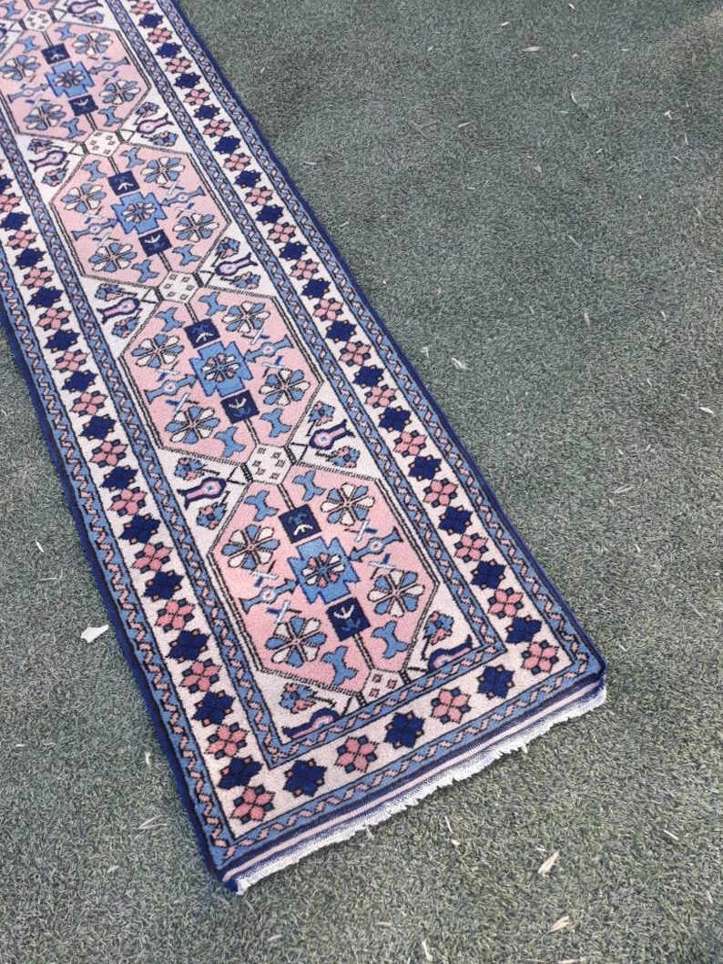 May include: A long, narrow rug with a pink, blue, and white geometric pattern. The rug has a fringed edge and is made of wool.