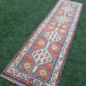 May include: A long, narrow rug with a geometric pattern in shades of pink, blue, green, and white. The rug has a fringe border.