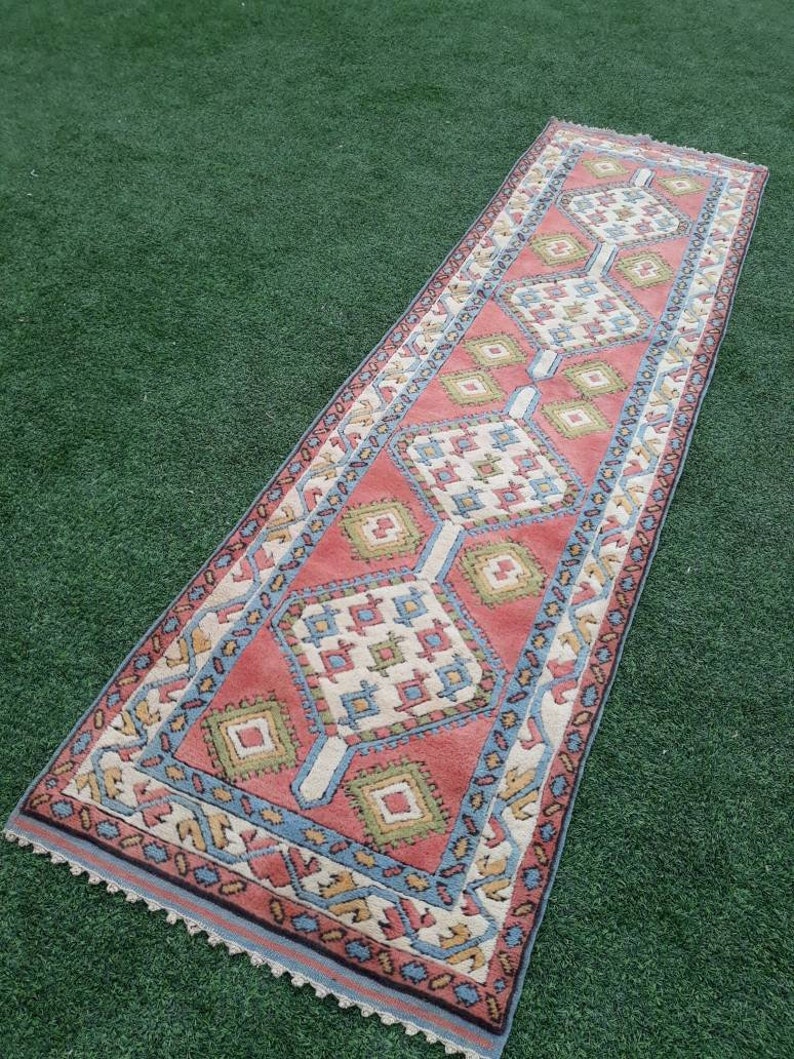 May include: A long, narrow rug with a geometric pattern in shades of pink, blue, green, and white. The rug has a fringe border.