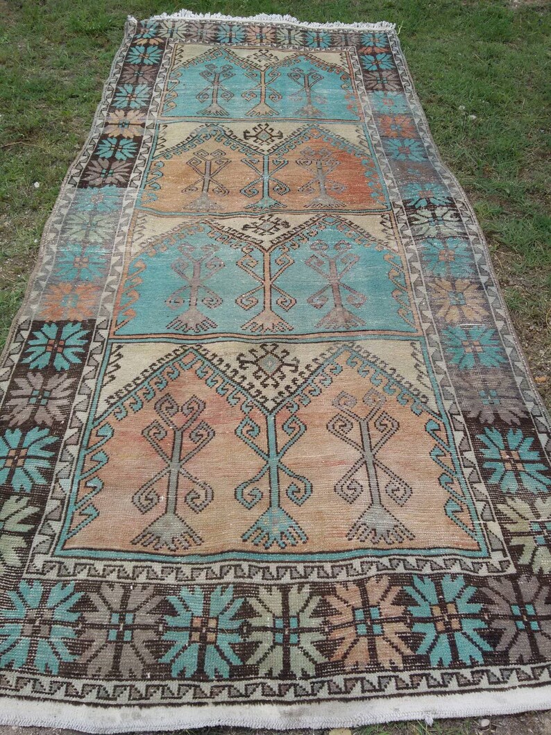 Puede incluir: Una alfombra turca vintage con un patr&oacute;n geom&eacute;trico en tonos azules, marrones y beige. La alfombra tiene un aspecto descolorido y desgastado y presenta un motivo de &aacute;rbol repetido.