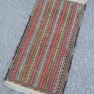 May include: A long, narrow rug with a geometric pattern in shades of red, green, brown, and white. The rug has a fringe border.