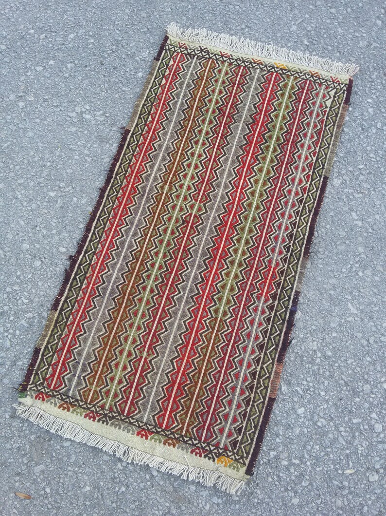 May include: A long, narrow rug with a geometric pattern in shades of red, green, brown, and white. The rug has a fringe border.