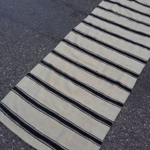 Pu&ograve; includere: Un tappeto lungo e bianco con strisce nere. Le strisce sono realizzate in un materiale spesso e tessuto e hanno un aspetto leggermente strutturato. Il tappeto &egrave; steso su una superficie di asfalto grigia.