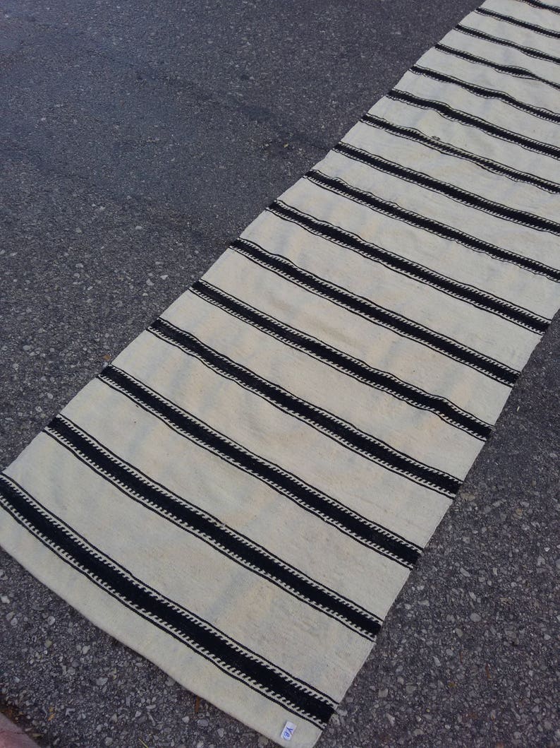 Pu&ograve; includere: Un tappeto lungo e bianco con strisce nere. Le strisce sono realizzate in un materiale spesso e tessuto e hanno un aspetto leggermente strutturato. Il tappeto &egrave; steso su una superficie di asfalto grigia.