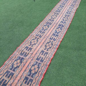 May include: A long, narrow rug with a pink, blue, and white geometric pattern. The rug is laid out on a green surface.